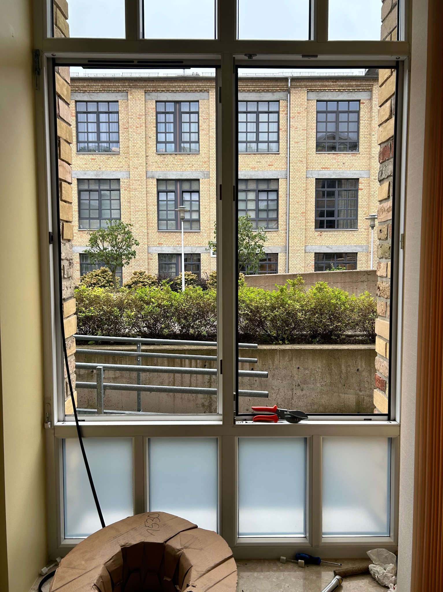 Widok z wnętrza na dwuskrzydłowe okno z szybami częściowo matowymi, z narzędziami leżącymi na parapecie i kartonowym elementem na podłodze, na zewnątrz budynek z cegły i zieleń.