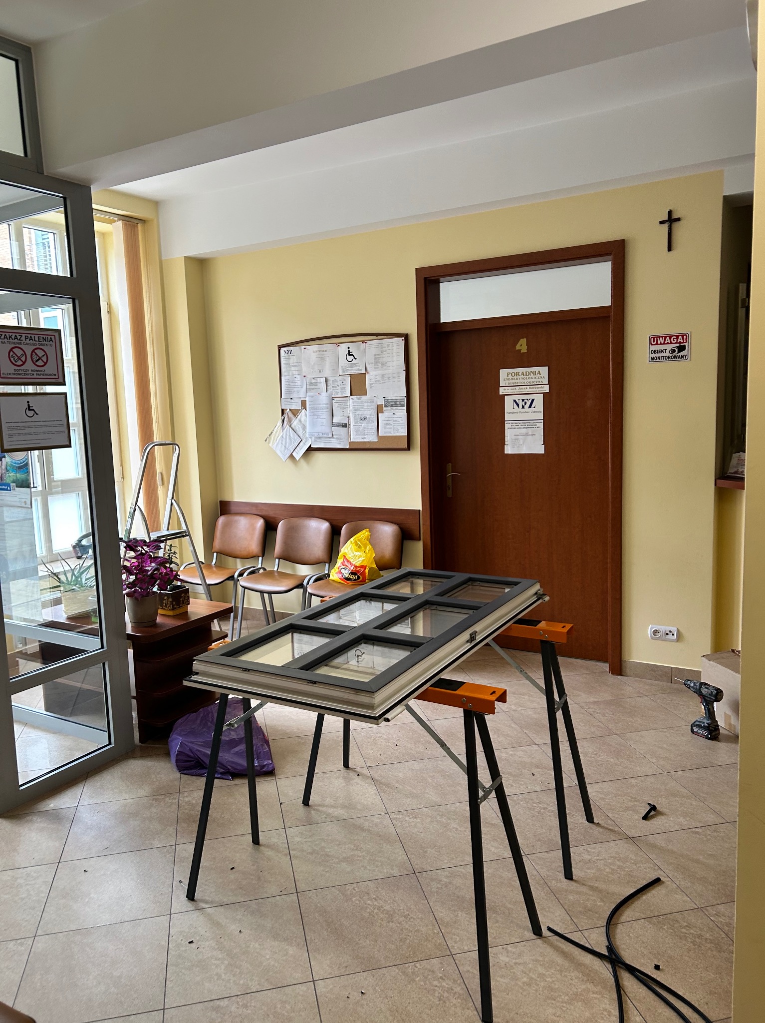 Demontaż okna z sześcioma szybami, o szarej ramie, leżącego na kozłach w poczekalni przychodni, widoczna wiertarka akumulatorowa, listwy uszczelniające oraz drabina.