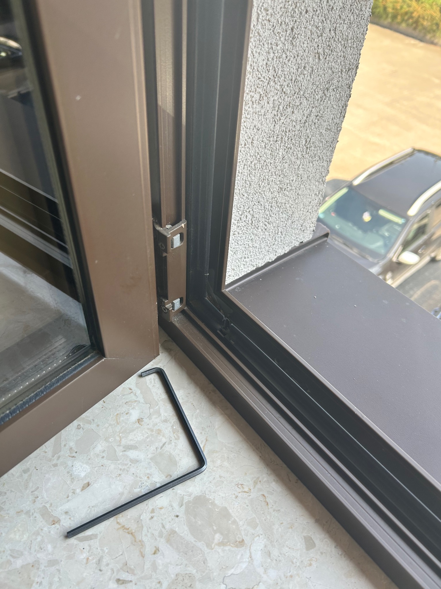 Brązowe okno PCV z widocznym mechanizmem regulacji i kluczem imbusowym leżącym na marmurowym parapecie podczas regulacji.