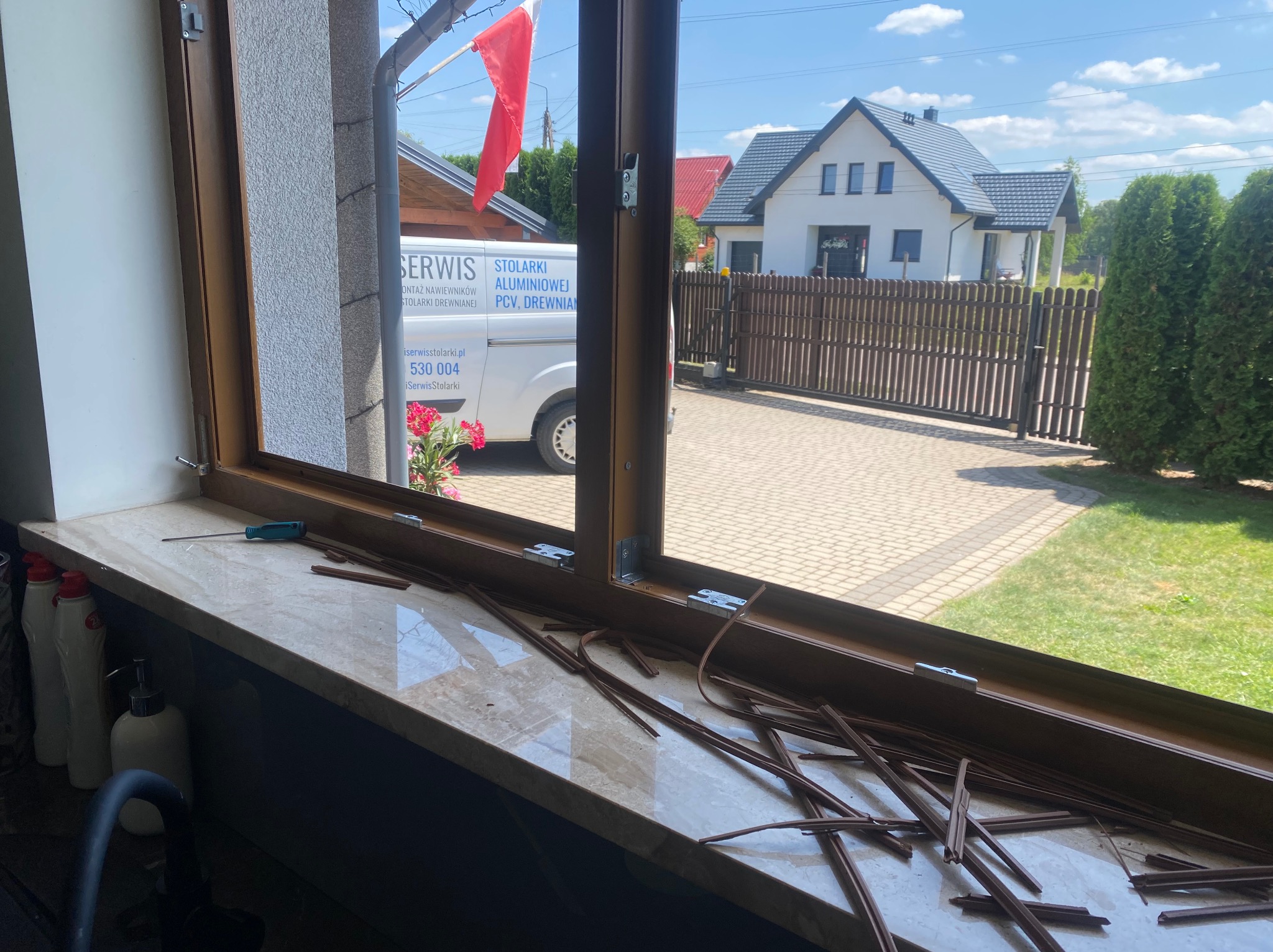 Brązowe uszczelki okienne leżące na marmurowym parapecie podczas montażu, na zewnątrz widoczny samochód serwisowy z polską flagą i logo firmy zajmującej się stolarką okienną.