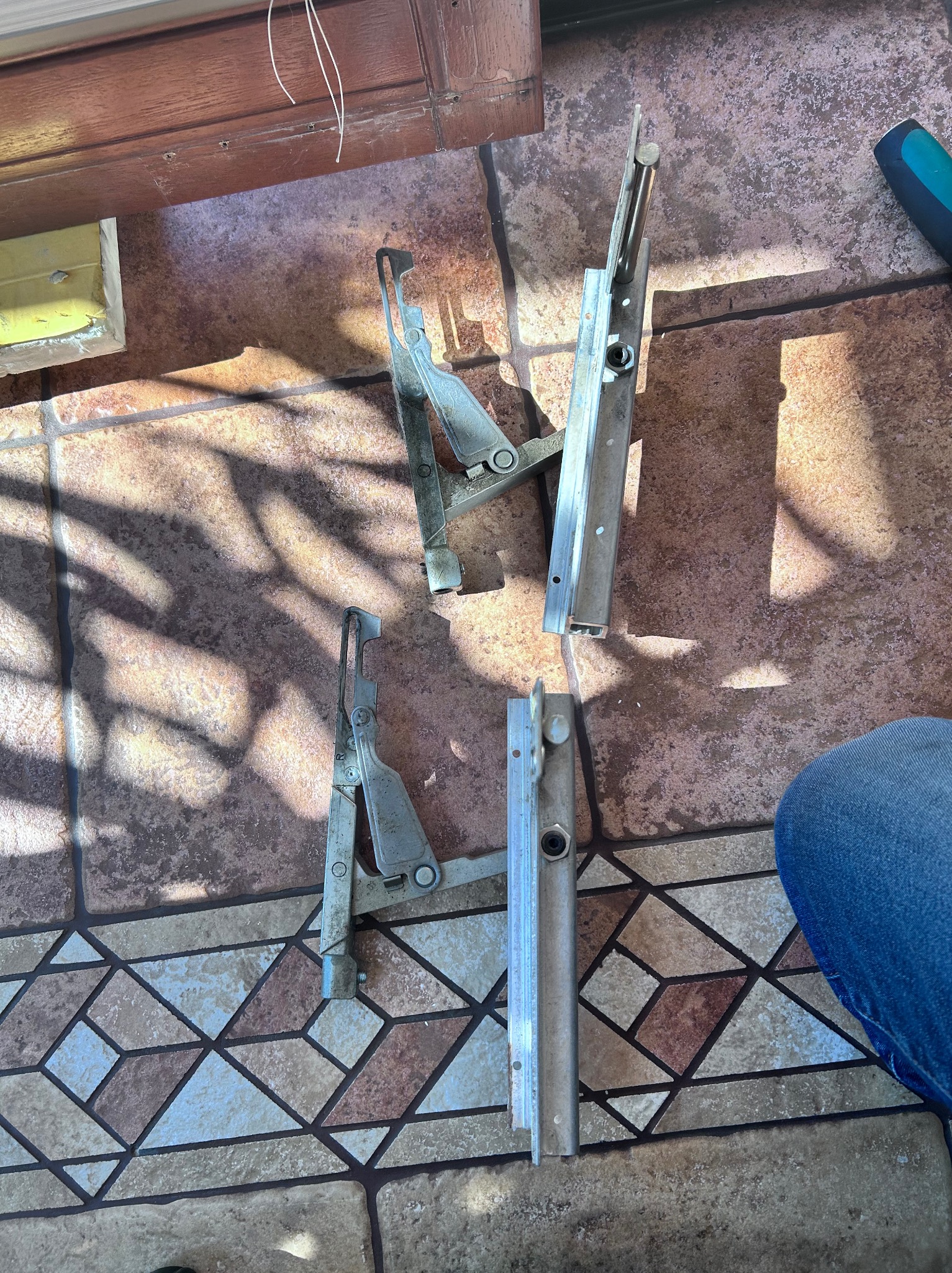 Dwa komplety srebrnych okuć okiennych leżące na podłodze z płytek w geometryczny wzór, obok widoczny fragment niebieskich dżinsów.