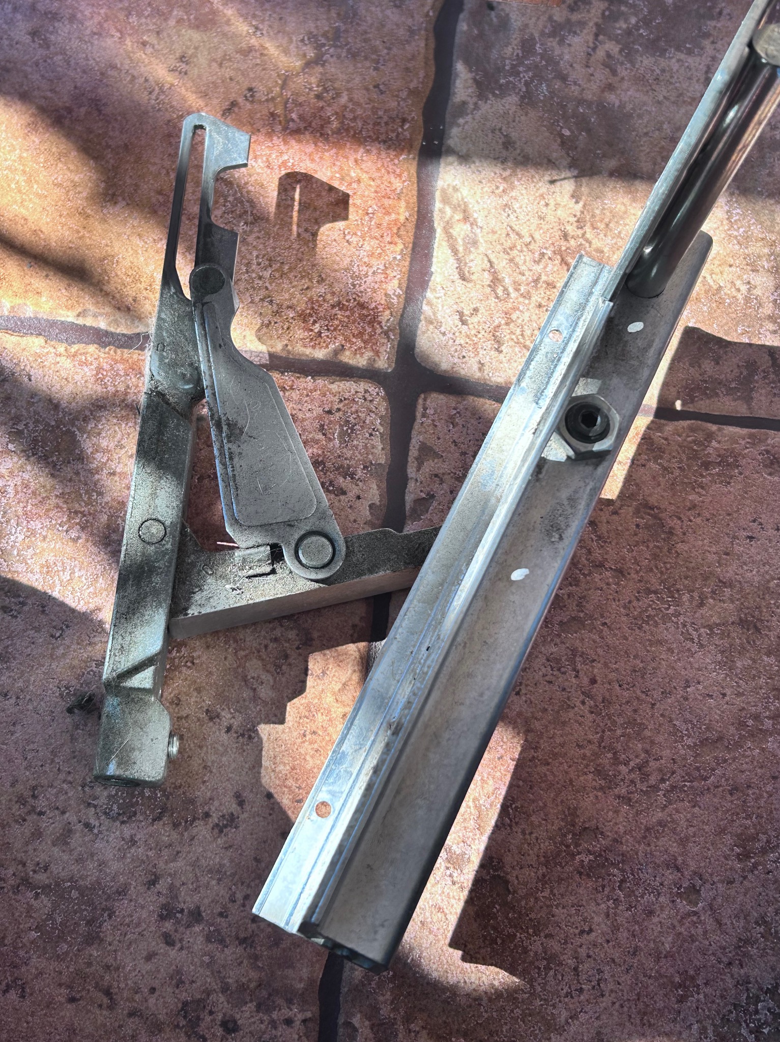 Metalowe elementy okienne, prawdopodobnie mechanizm otwierania i fragment ramy, leżące na brązowej powierzchni przypominającej płytki lub kamień, oświetlone ostrym światłem słonecznym.