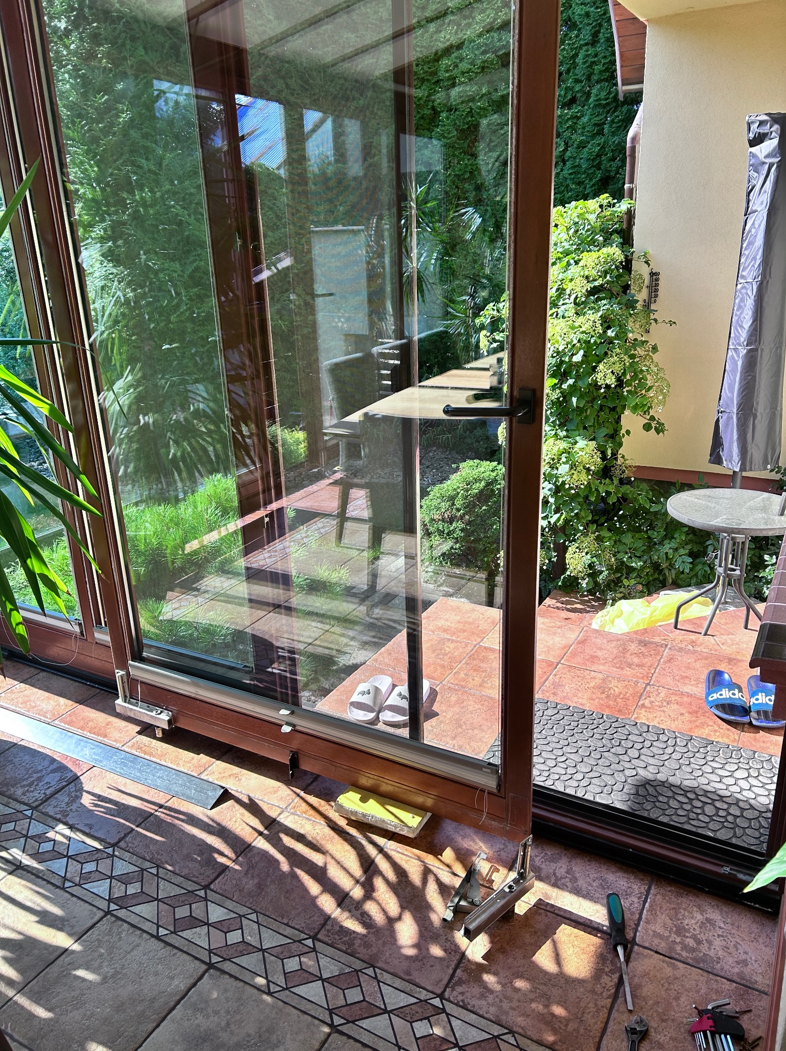 Otwarta przesuwna szyba z brązowymi ramami, widoczne narzędzia i akcesoria do montażu na kafelkach podłogowych, zieleń ogrodu widoczna przez szybę.