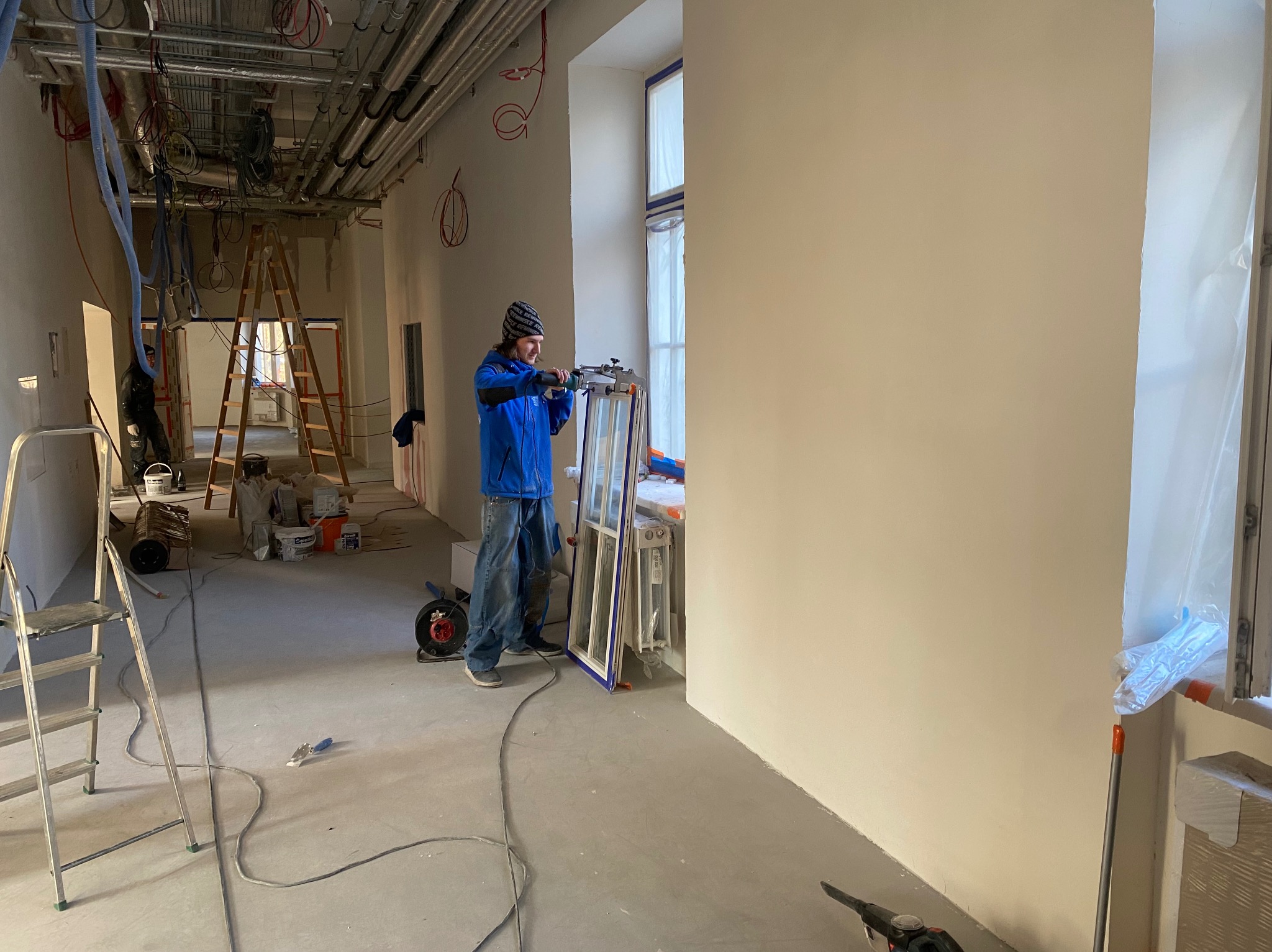 Mężczyzna w niebieskiej kurtce montuje okno w nowym budynku, widoczne instalacje na suficie i drabiny