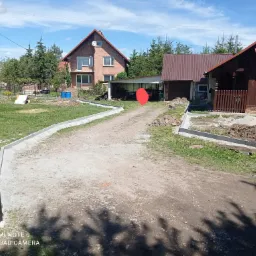 Niedokończona ścieżka z betonowych obrzeży prowadząca do domu z cegły i garażu, widok na posesję w słoneczny dzień.