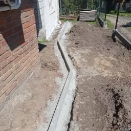 Wykopany rów z betonowym obrzeżem wzdłuż ściany budynku z cegły i białego bloczka, widoczne korzenie w ziemi, w tle ogrodzenie z siatki i sterta płyt.