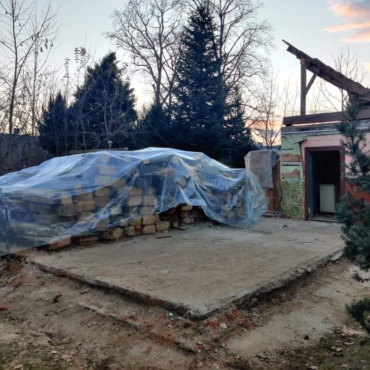 Przygotowanie do prac brukarskich: stos cegieł przykryty folią na przygotowanym podłożu pod taras, w tle częściowo rozebrany budynek.