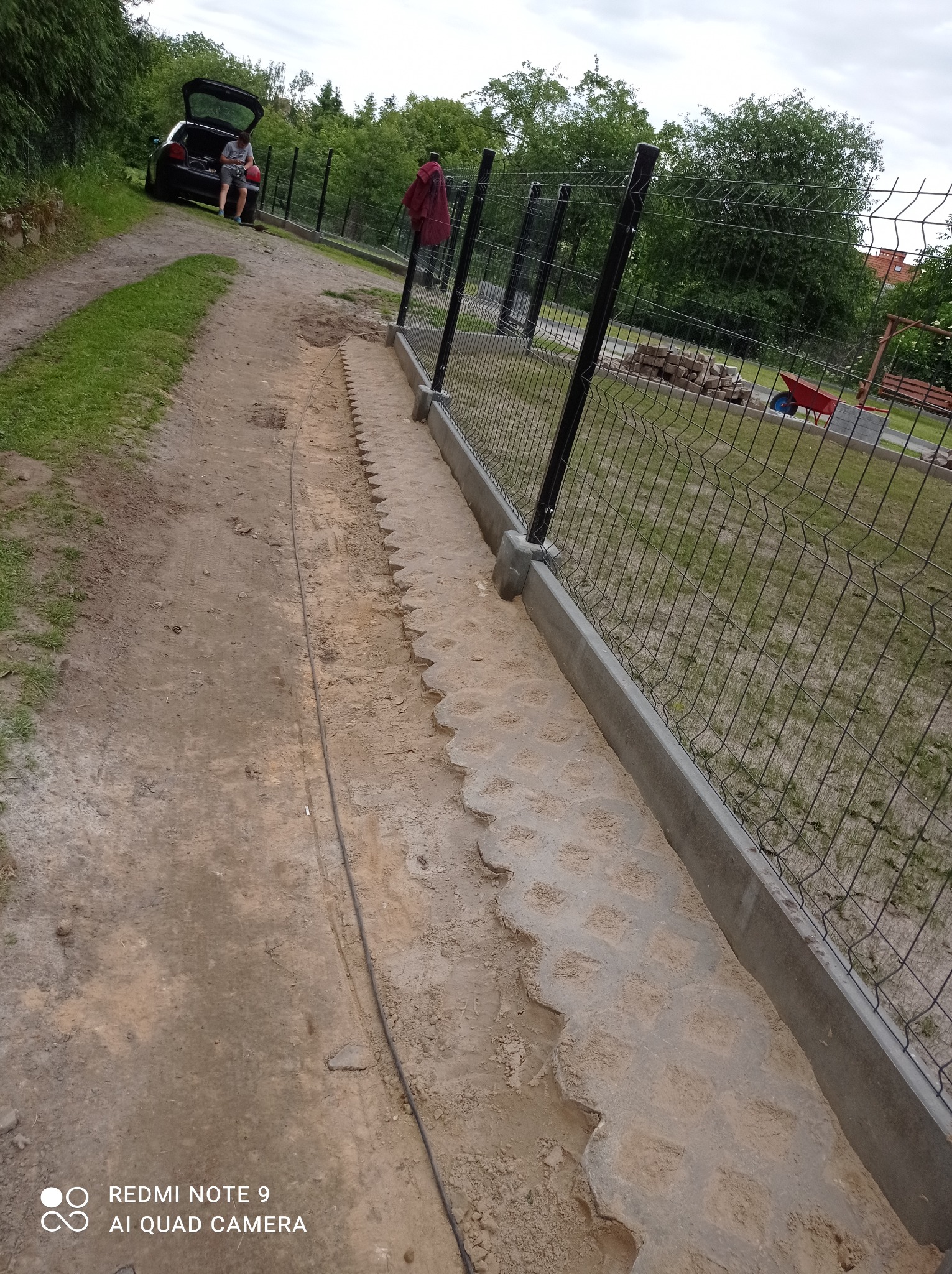 Przygotowanie podłoża pod ogrodzenie z betonową obrzeżem, widoczne wyprofilowane koryto w ziemi z odciśniętymi wzorami, siatka ogrodzeniowa w tle, osoba przy otwartym bagażniku samochodu.