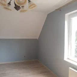 Pokój po odświeżeniu, ściany pomalowane na szaro, jasna podłoga, ozdobny żyrandol i karnisz.