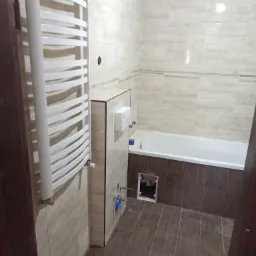 Wykończona łazienka z beżowymi płytkami imitującymi marmur, brązowymi płytkami pod wanną, białym grzejnikiem drabinkowym i otworem rewizyjnym w zabudowie wanny.