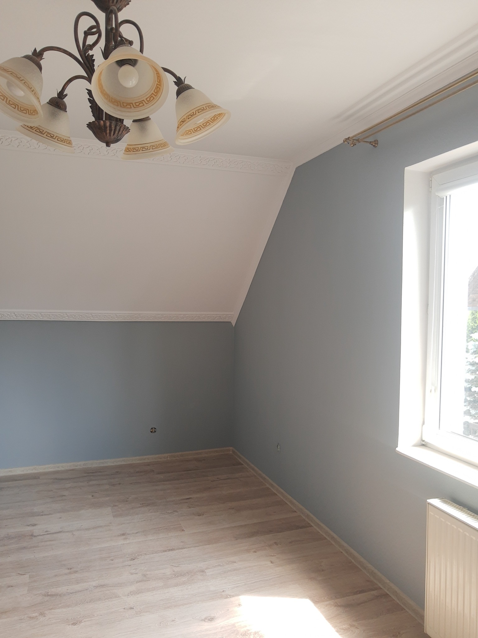 Pokój po odświeżeniu, ściany pomalowane na szaro, jasna podłoga, ozdobny żyrandol i karnisz.