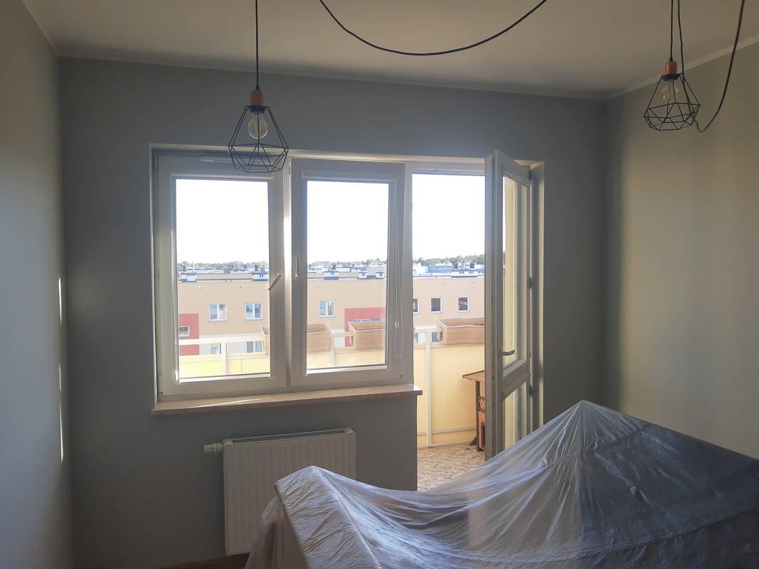 Szare ściany w pokoju przed odświeżeniem, okno balkonowe z widokiem na osiedle, meble zabezpieczone folią, wiszące lampy druciane.