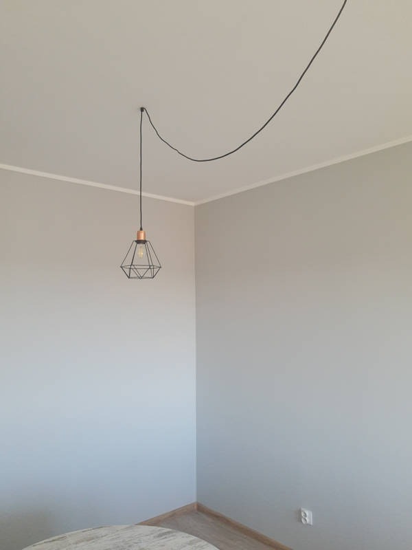 Pokój z szarymi ścianami, białym sufitem i listwami przypodłogowymi, z wiszącą lampą drucianą w kształcie diamentu z miedzianym elementem i czarnym kablem.
