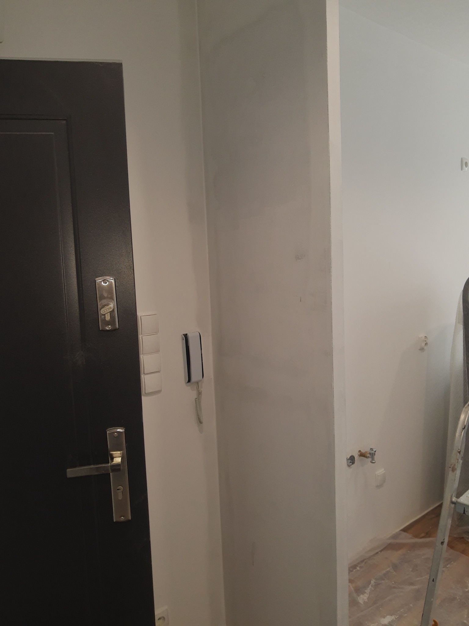 Przygotowana ściana pod malowanie, widoczne ślady szpachlowania i zatarte miejsca, drzwi wejściowe w ciemnym kolorze po lewej stronie, zabezpieczona podłoga folią malarską, drabina malarska w tle.