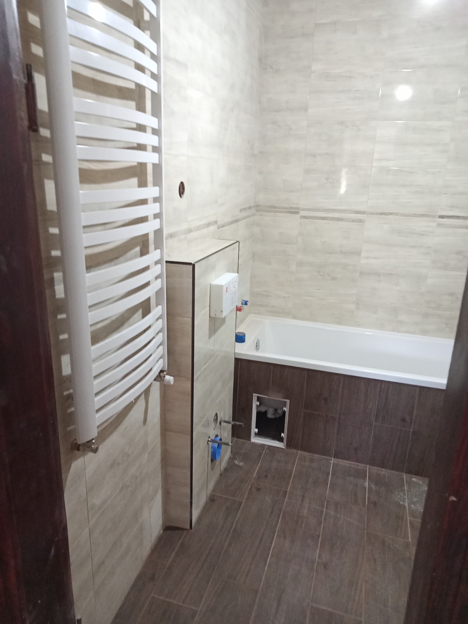 Wykończona łazienka z beżowymi płytkami imitującymi marmur, brązowymi płytkami pod wanną, białym grzejnikiem drabinkowym i otworem rewizyjnym w zabudowie wanny.