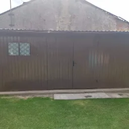 Brązowy garaż blaszany z oknem z wzorzystą folią, widoczny na tle trawnika i starego budynku.