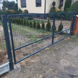 Nowo zamontowana, grafitowa brama panelowa z widocznymi elementami mocującymi, osadzona na betonowej podmurówce, na tle domu z roletami zewnętrznymi i ozdobnymi krzewami.