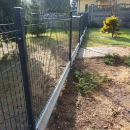 Długi, czarny, metalowy płot panelowy z betonową podmurówką oddzielający trawnik od posesji z żółtym domem w tle.