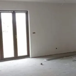 Wnętrze pomieszczenia w stanie deweloperskim, świeżo po nałożeniu gładzi gipsowej na ścianach i wylewce posadzki, widoczne aluminiowe profile podłogowe i wystające przewody elektryczne z sufitu.