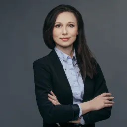 Portret kobiety z Krakowa w ciemnej marynarce i błękitnej koszuli w drobne wzory, z założonymi rękami na tle szarego studia.