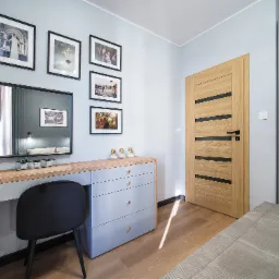 Jasny pokój z drewnianą podłogą, szarą komodą z lustrem i czarnym krzesłem. Na ścianie wiszą czarno-białe zdjęcia w ramkach. W tle widoczna drewniana wewnętrzna ościeżnica z czarnymi akcentami.