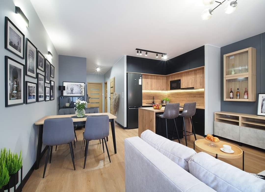 Jasne, przestronne wnętrze mieszkania z aneksem kuchennym, drewnianymi meblami, szarymi ścianami i czarnymi akcentami, oświetlone punktowo i listwami LED, z widoczną galerią czarno-białych zdjęć...