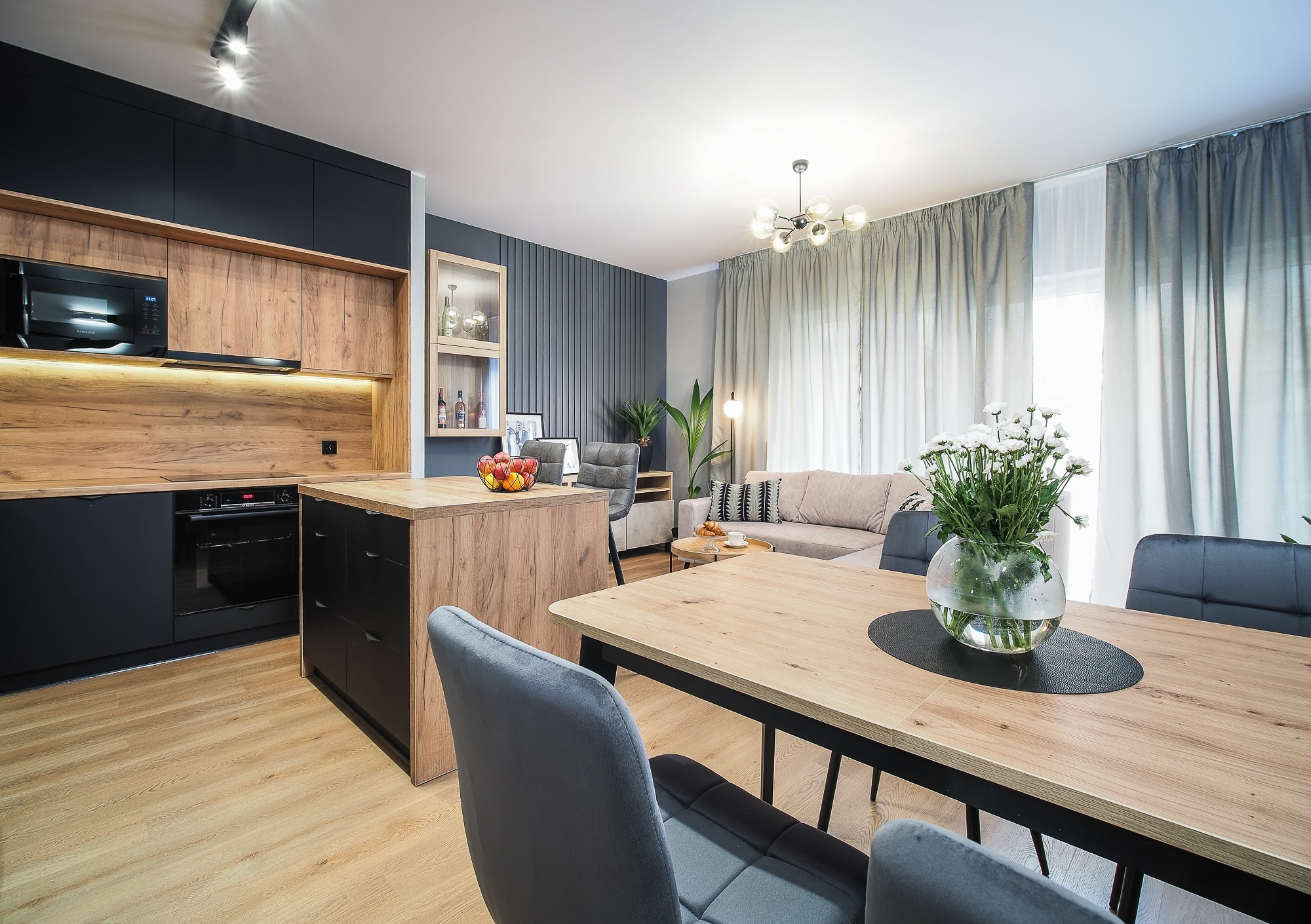 Nowoczesne wnętrze: czarna zabudowa kuchenna z drewnianymi akcentami, wyspa kuchenna, stół z bukietem białych kwiatów i szarymi krzesłami, w tle salon z beżową sofą i dekoracyjną ścianą.