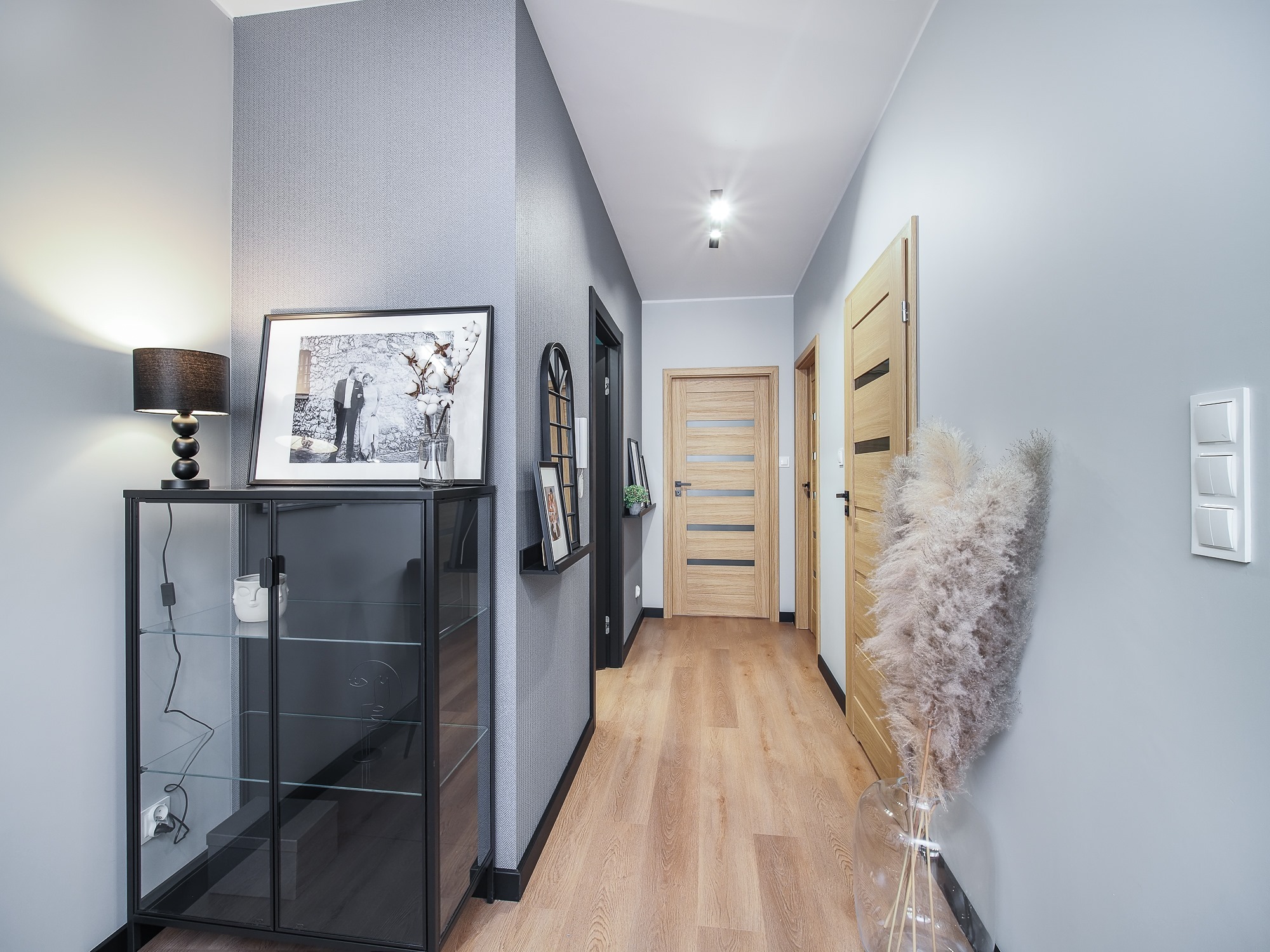 Nowoczesny przedpokój z drewnianą podłogą, szarą tapetą w jodełkę, szklaną witryną z dekoracjami i dwiema drewnianymi parami drzwi z czarnymi ościeżnicami.