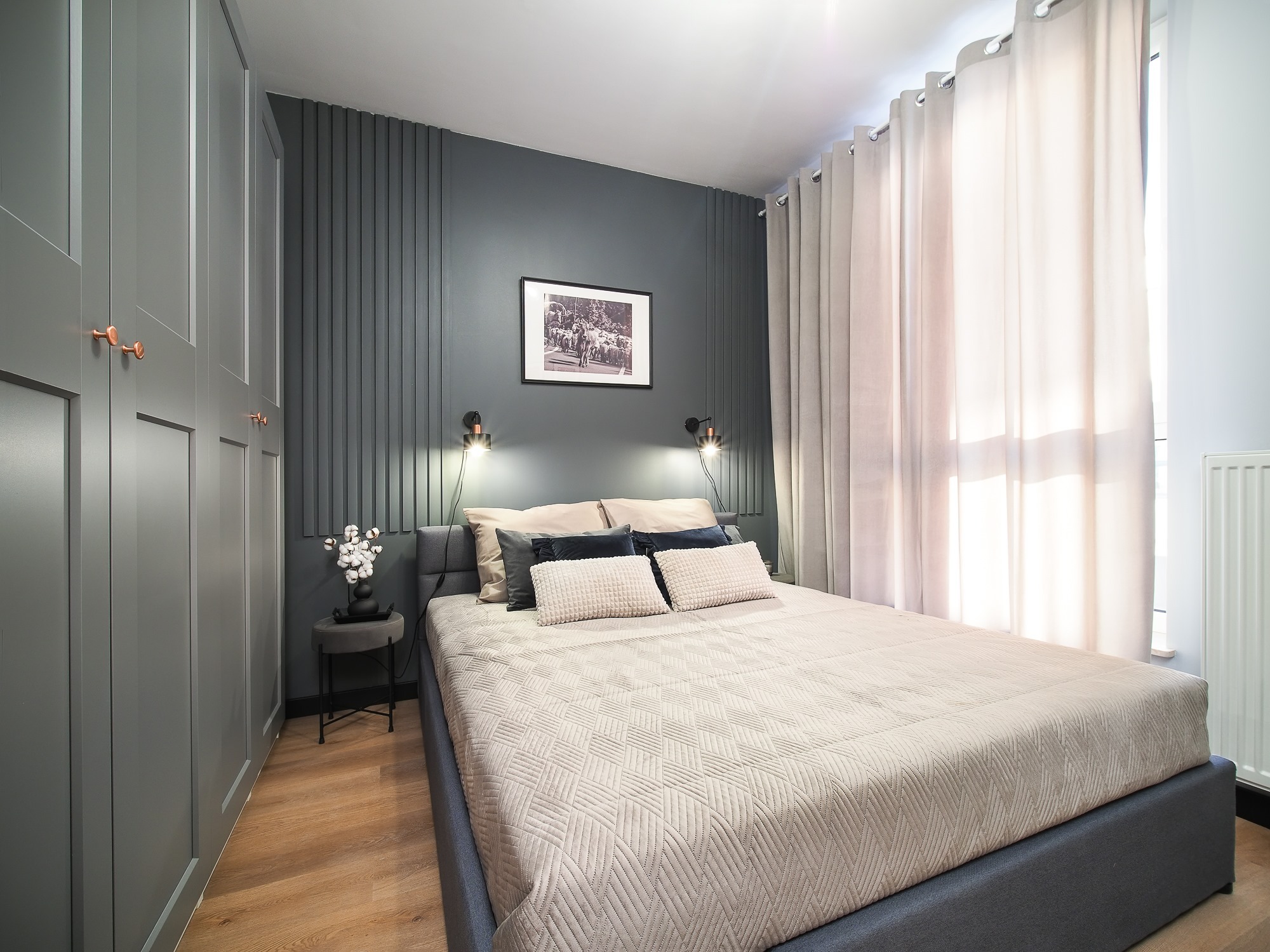 Nowoczesna sypialnia z szarą tapetą z pionowymi panelami, łóżkiem z beżową narzutą, szarą szafą z miedzianymi uchwytami i drewnianą podłogą.