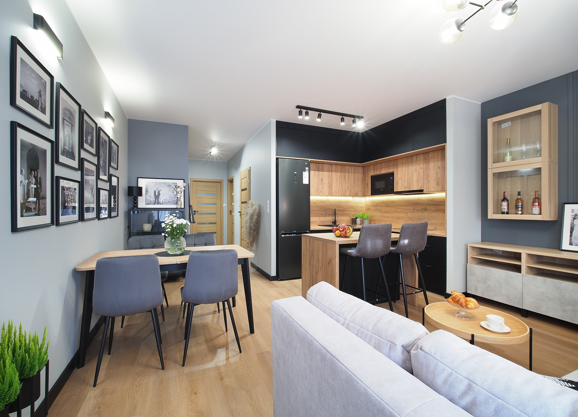 Jasne, przestronne wnętrze mieszkania z drewnianą podłogą, szarymi ścianami i nowoczesną kuchnią z drewnianymi szafkami, czarnymi akcentami i barkiem z hokerami. W części jadalnej stół z szarymi...