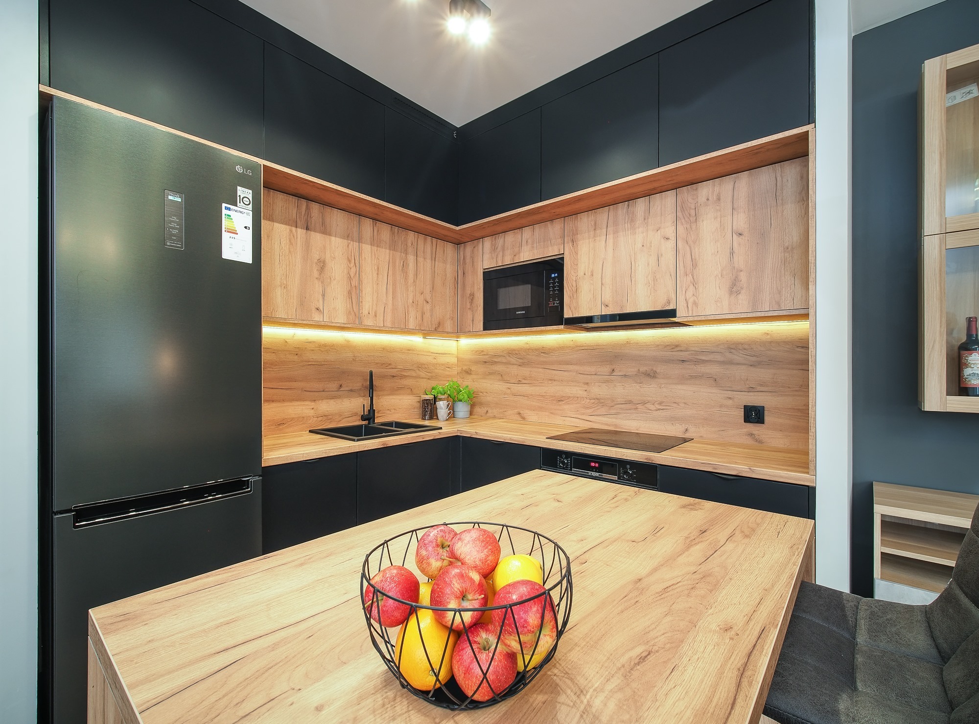 Nowoczesna kuchnia z drewnianymi frontami szafek i czarnymi elementami, oświetleniem LED pod szafkami górnymi, czarnym zlewem i baterią, oraz koszem z owocami na wyspie kuchennej.