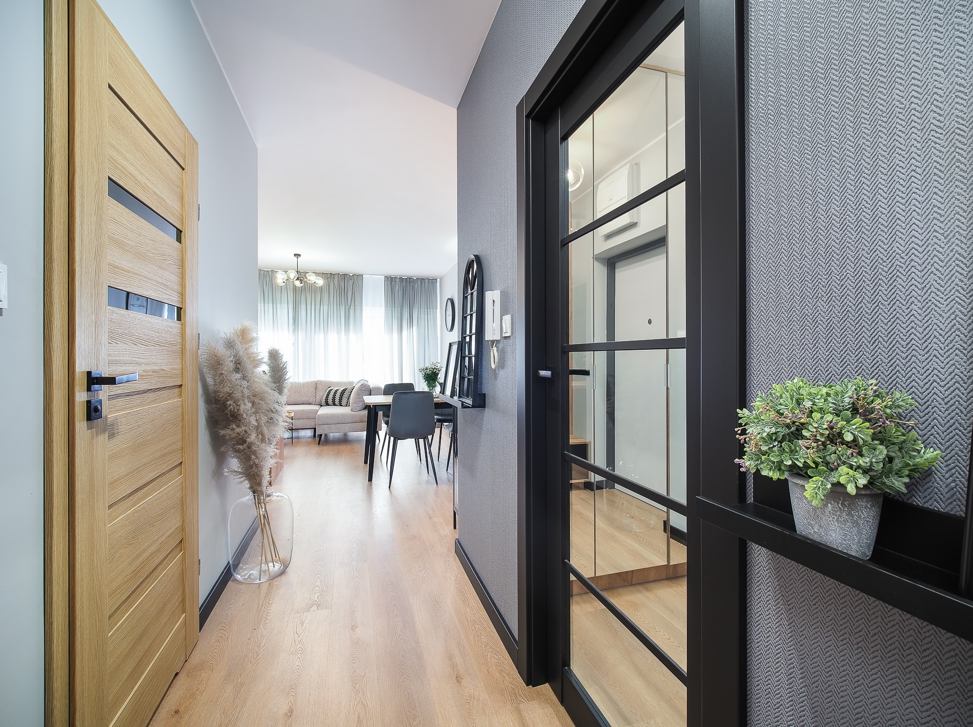 Nowoczesne wnętrze mieszkania: dębowa podłoga, szara tapeta w jodełkę, drzwi z czarnymi szprosami, drewniane drzwi z przeszkleniami, dekoracyjny bukiet w wazonie i roślina doniczkowa na półce.