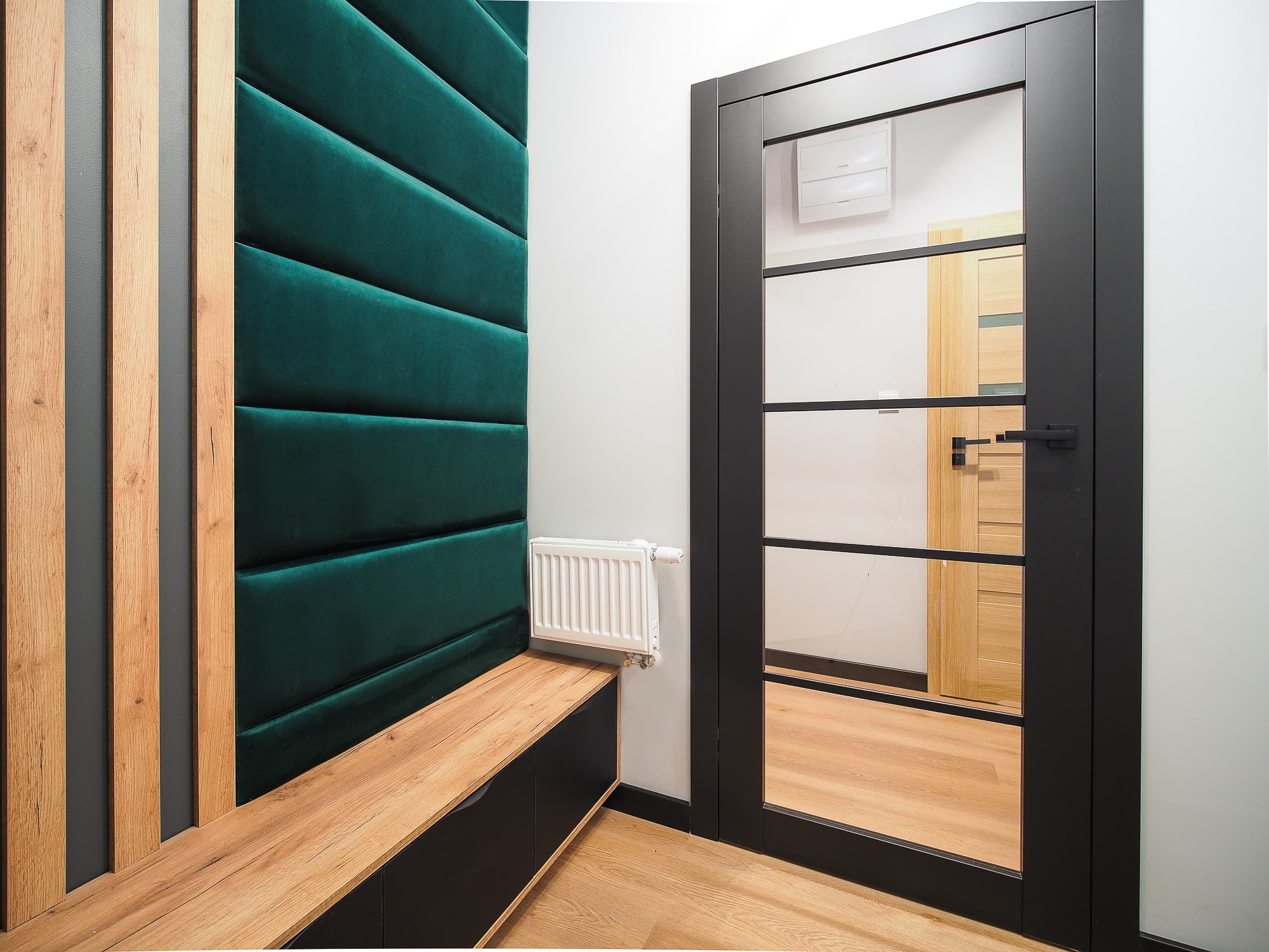 Nowoczesny przedpokój z panelem tapicerowanym w kolorze butelkowej zieleni, drewnianą zabudową, grzejnikiem i czarnymi drzwiami z przeszkleniami.
