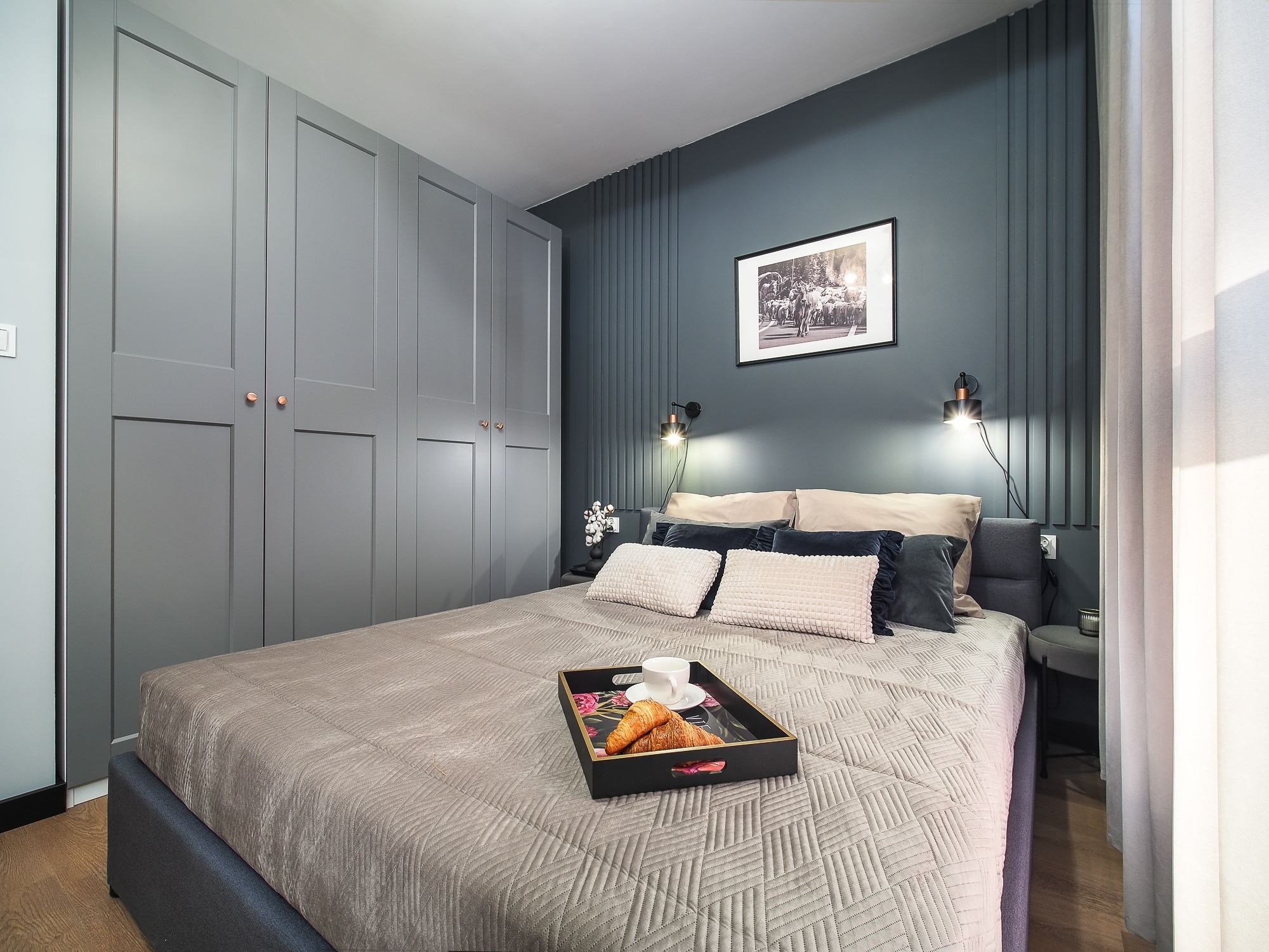 Nowoczesna sypialnia z szarą zabudową szaf, panelem ściennym w pionowe pasy, łóżkiem z szarą narzutą i śniadaniem na tacy, oświetlona lampkami nocnymi.