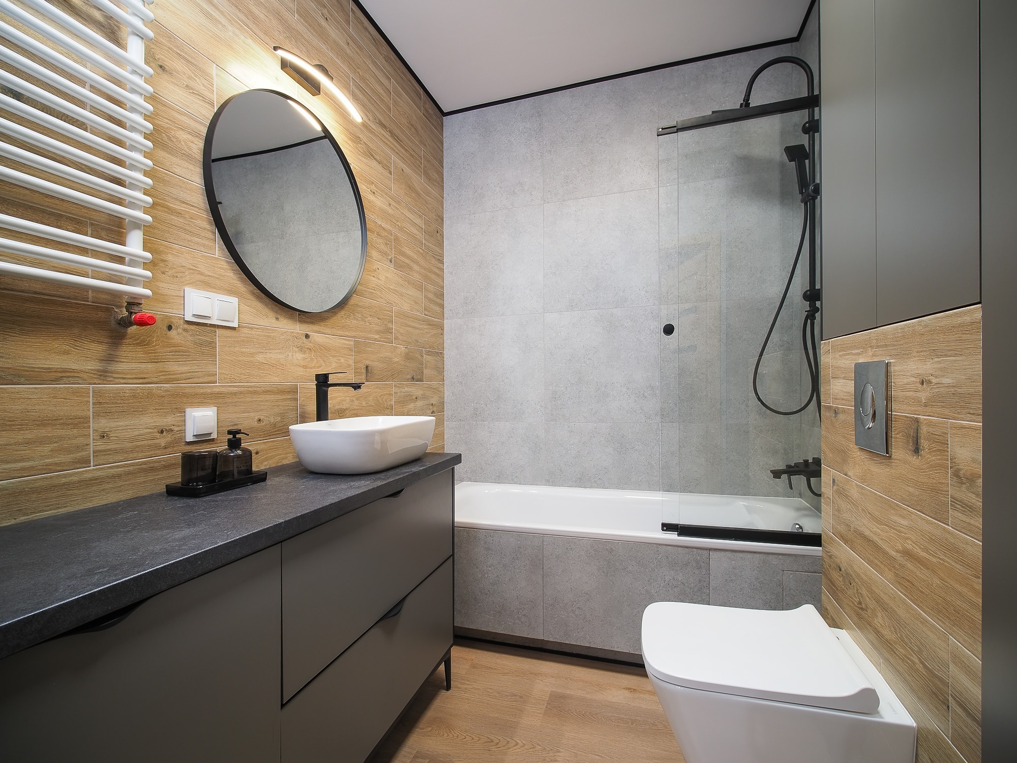 Nowoczesna łazienka z drewnopodobnymi płytkami na ścianach, szarą zabudową umywalki z czarnym blatem, owalnym lustrem z podświetleniem, białą umywalką nablatową, czarną armaturą, grzejnikiem...