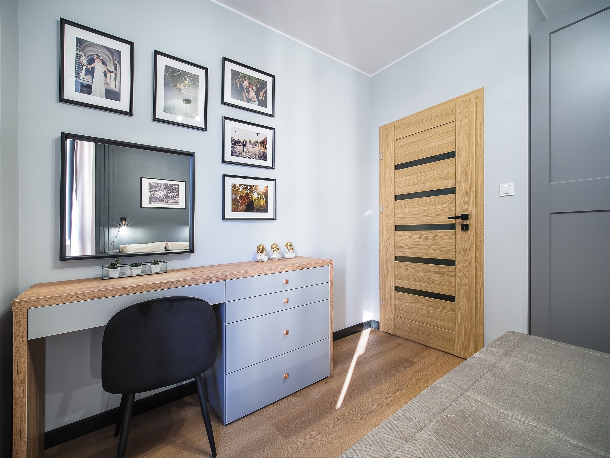 Jasny pokój z drewnianą podłogą, szarą komodą z lustrem i czarnym krzesłem. Na ścianie wiszą czarno-białe zdjęcia w ramkach. W tle widoczna drewniana wewnętrzna ościeżnica z czarnymi akcentami.