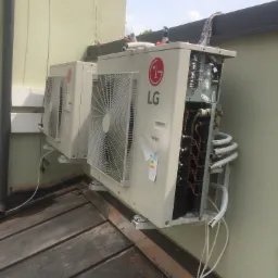 Dwa klimatyzatory LG zamontowane na zewnątrz budynku, jeden z otwartą obudową ukazującą wewnętrzne komponenty, widoczne rury i przewody.