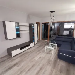 Nowoczesny salon z szarymi ścianami, meblami w kolorach bieli i grafitu, narożną sofą w odcieniach granatu oraz przeszklonymi drzwiami balkonowymi.