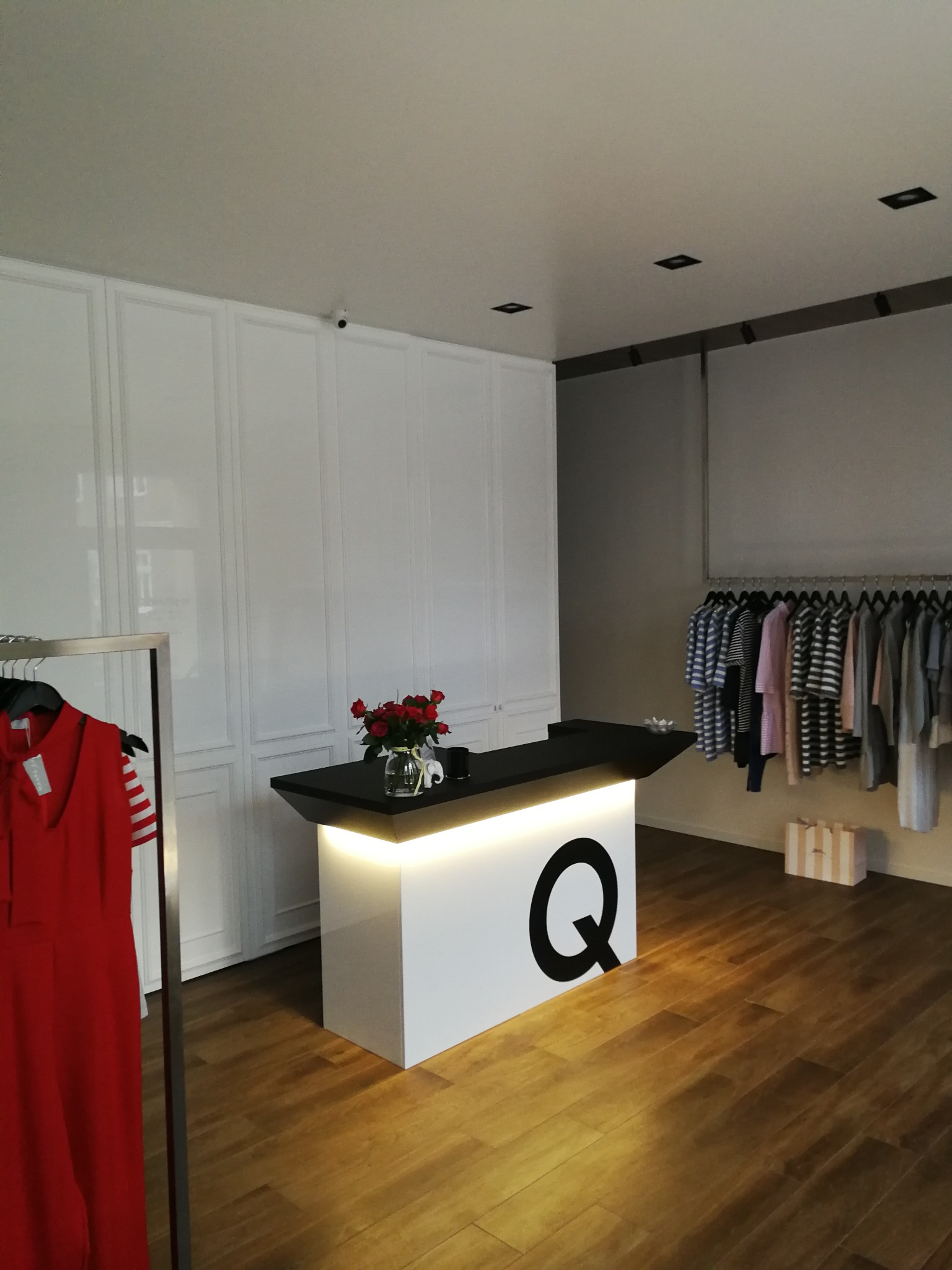 Wnętrze butiku z białymi szafami w zabudowie, ladą z podświetleniem LED i logiem 'Q', oraz wiszącymi ubraniami