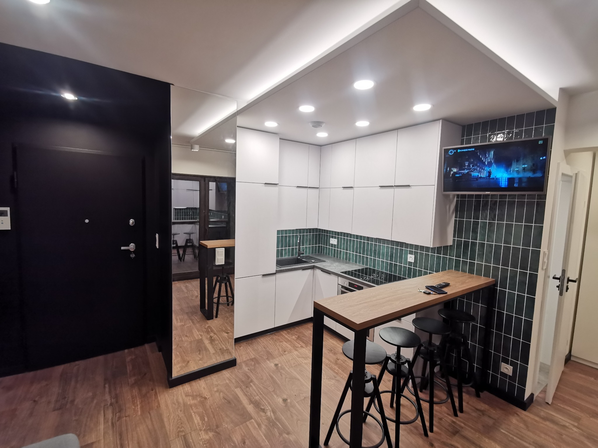Nowoczesna kuchnia z białymi szafkami, blatem barowym z hokerami, zielonymi płytkami na ścianie i telewizorem