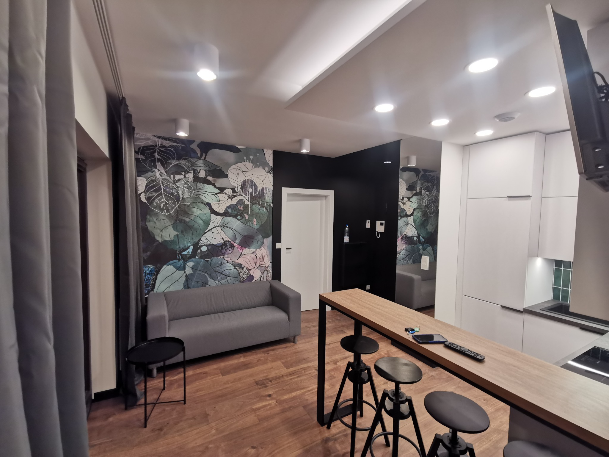 Nowoczesna kuchnia z białymi szafkami, drewnianym blatem barowym i czarnymi hokerami, sofa i tapeta w liście w tle.