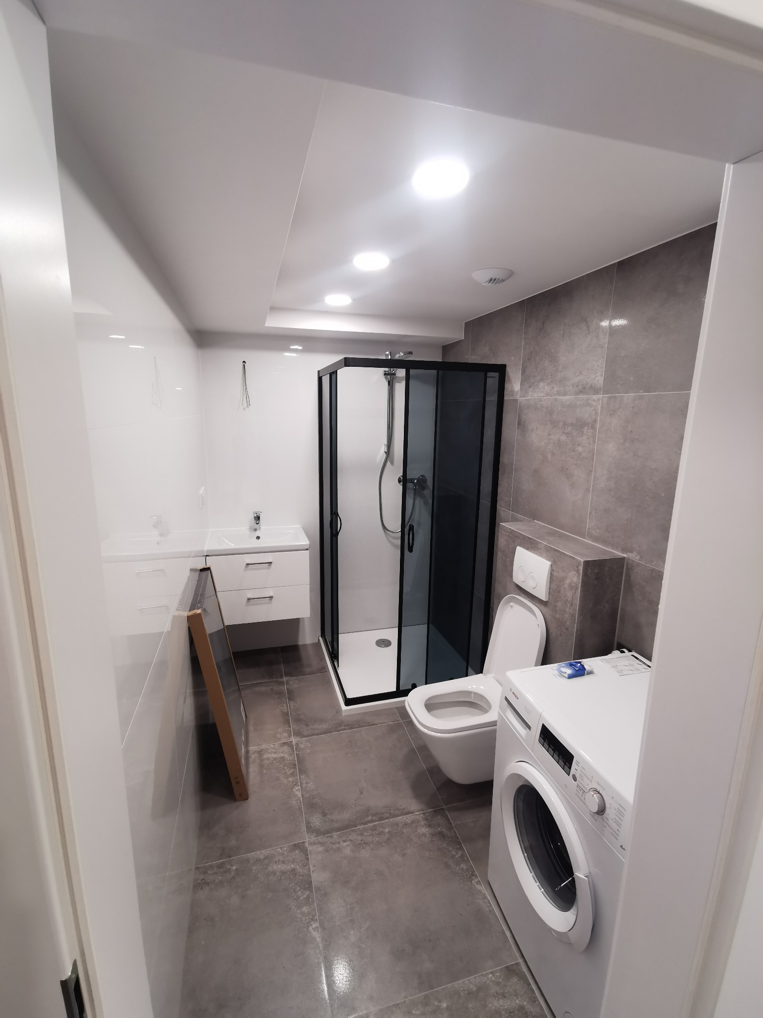 Nowoczesna łazienka z czarną kabiną prysznicową, białą umywalką z szafką, toaletą i pralką Bosch na szarych płytkach podłogowych, częściowo widoczna rama obrazu oparta o ścianę.