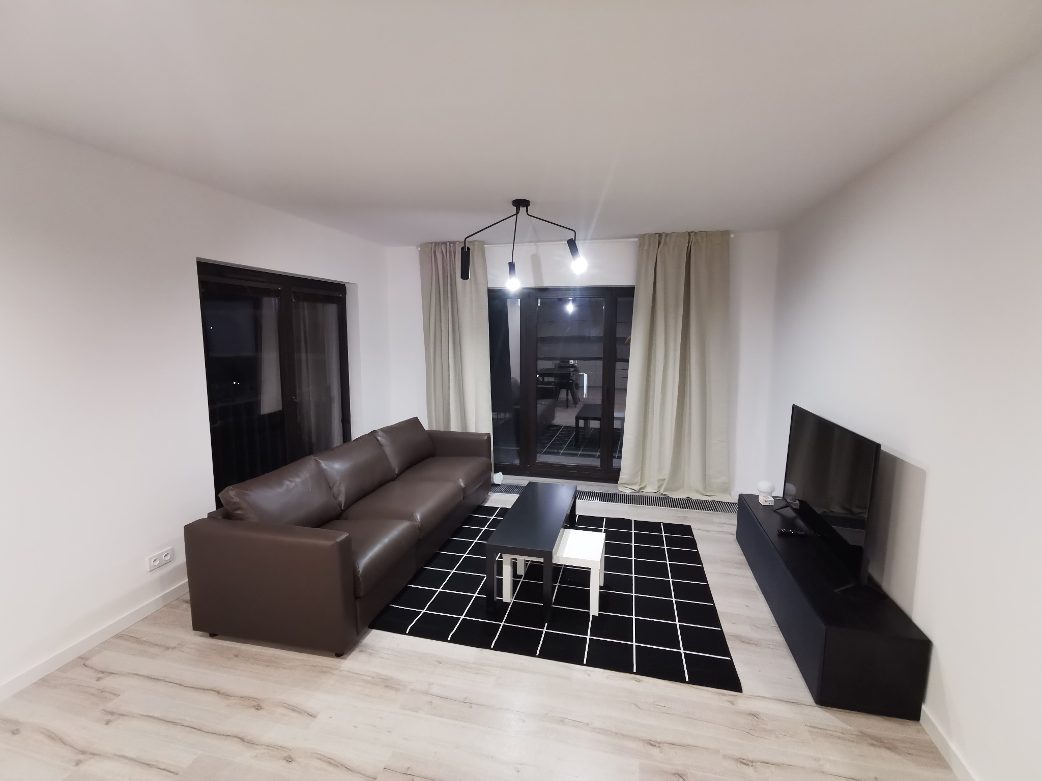 Nowoczesny salon z brązową skórzaną sofą, czarno-białym dywanem w kratę i czarną szafką RTV pod telewizorem, widok z szerokiego kąta.