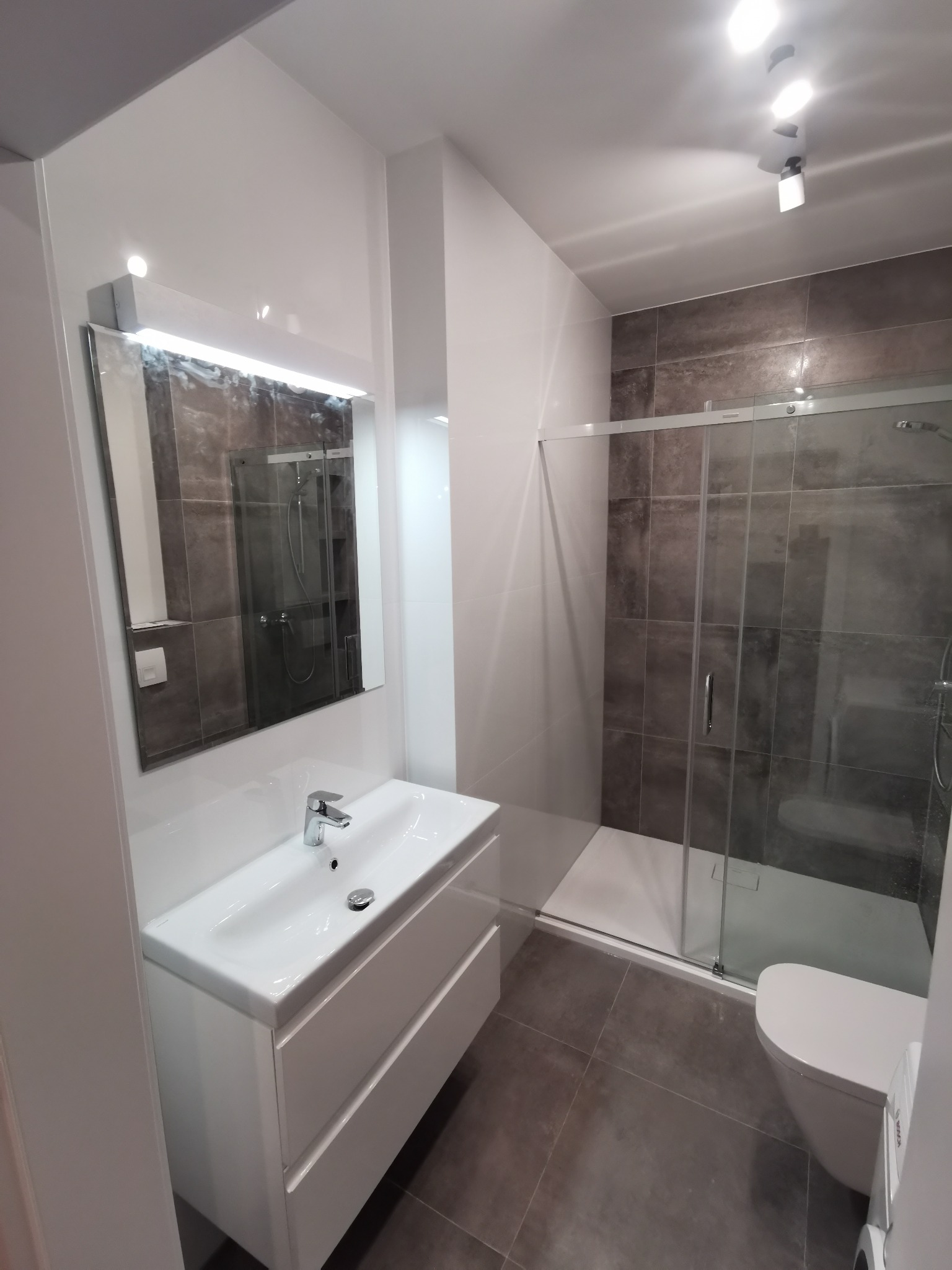 Nowoczesna łazienka z białymi meblami łazienkowymi, kabiną prysznicową z szarymi płytkami i oświetleniem punktowym