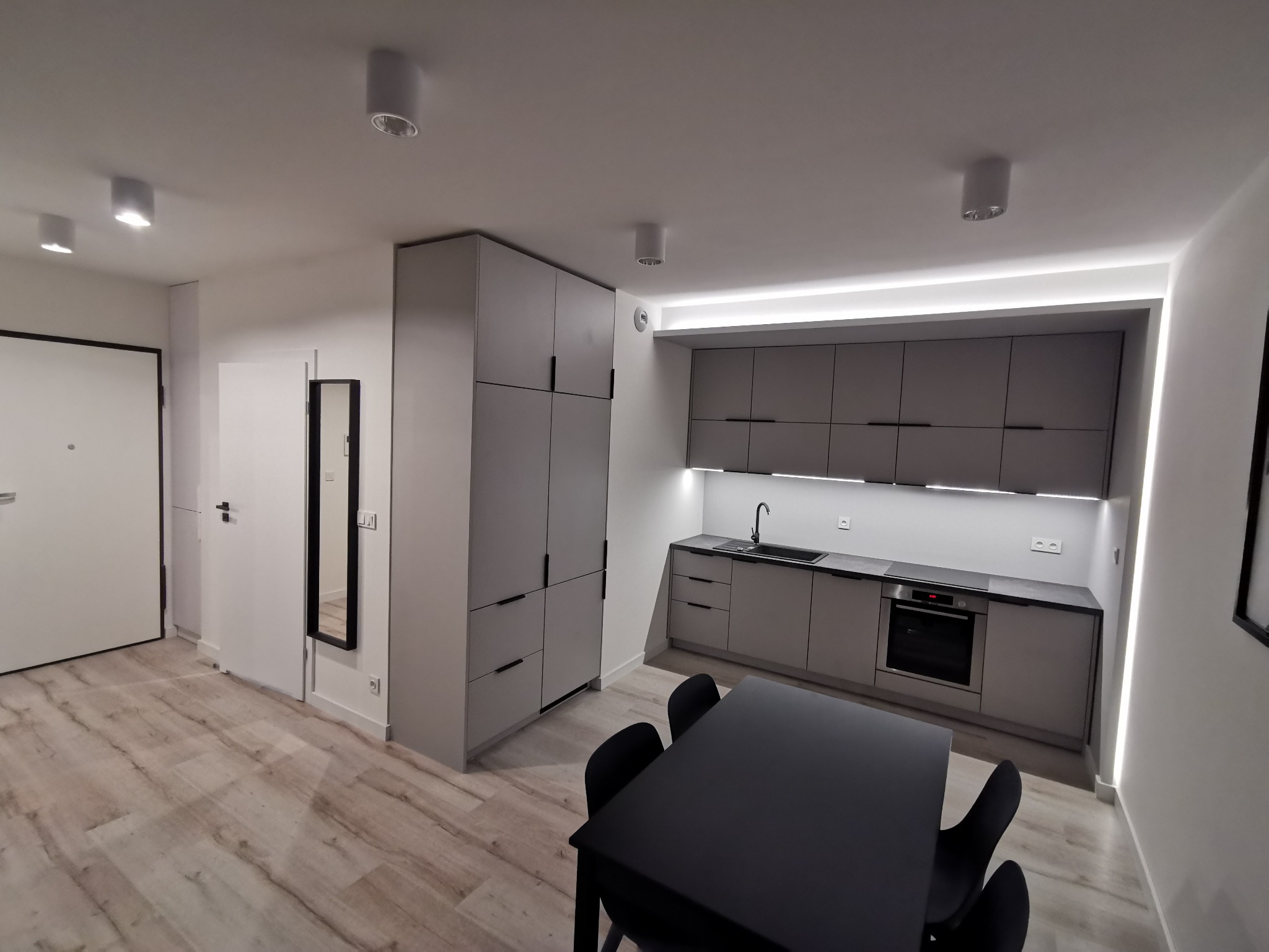 Nowoczesna kuchnia w odcieniach szarości z czarnym stołem i krzesłami, oświetleniem LED podszafkowym i pod sufitem, widok na przedpokój z lustrem.