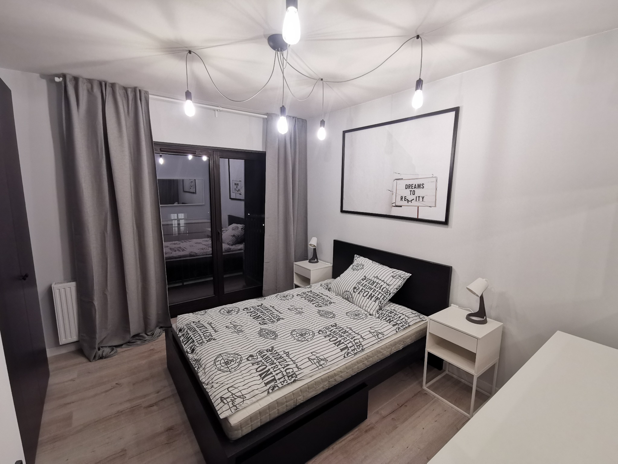Nowoczesna sypialnia z łóżkiem na czarnej ramie, pościel w paski z napisami, białe szafki nocne z lampkami, okno balkonowe z szarymi zasłonami i wiszące żarówki na kablach.