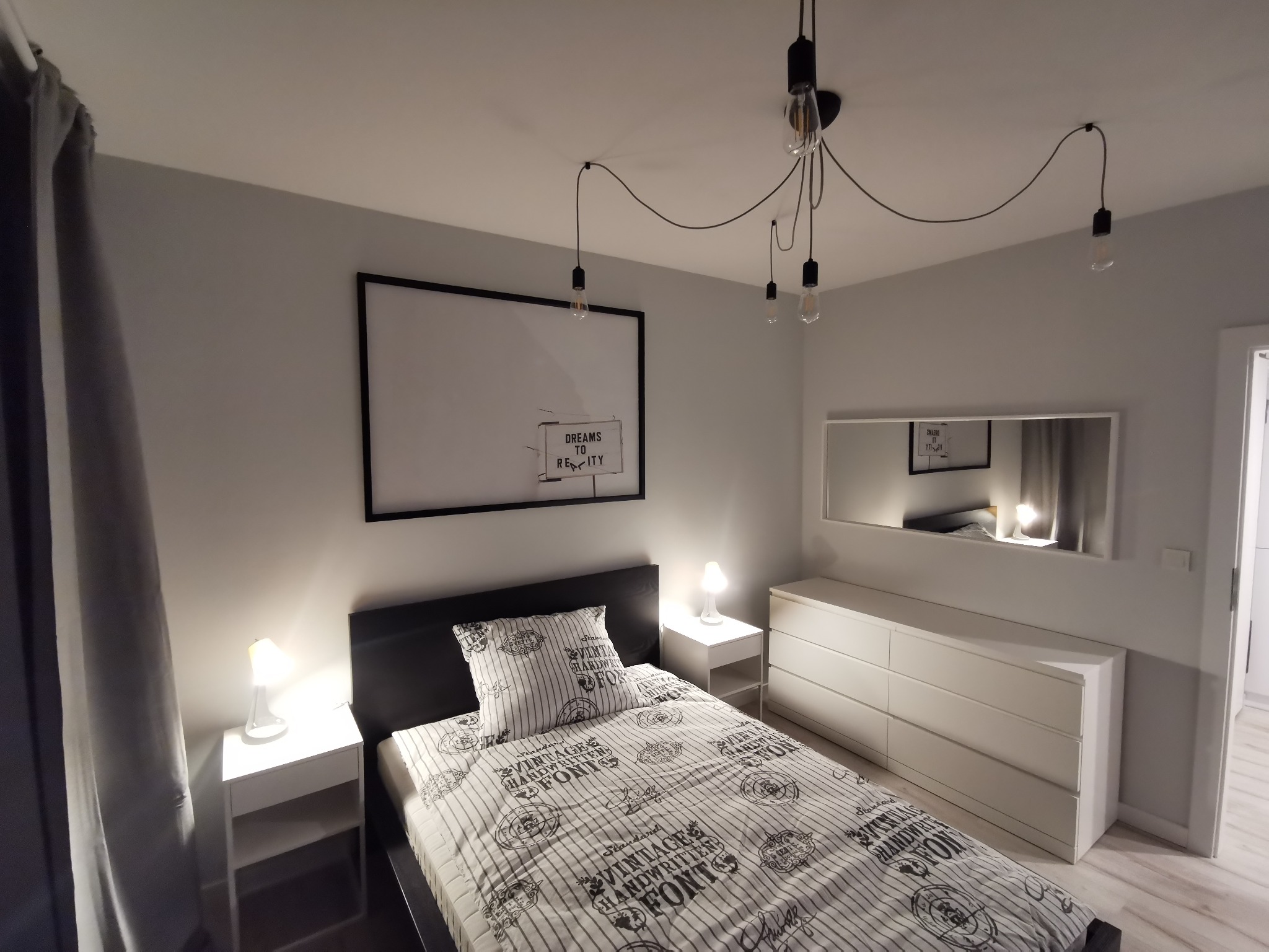 Nowoczesna sypialnia w Opolu z łóżkiem z czarną ramą i pościelą w paski, białą komodą i szafkami nocnymi z lampkami, oświetlona wiszącą lampą z żarówkami Edisona i ozdobiona plakatem w czarnej ramie.