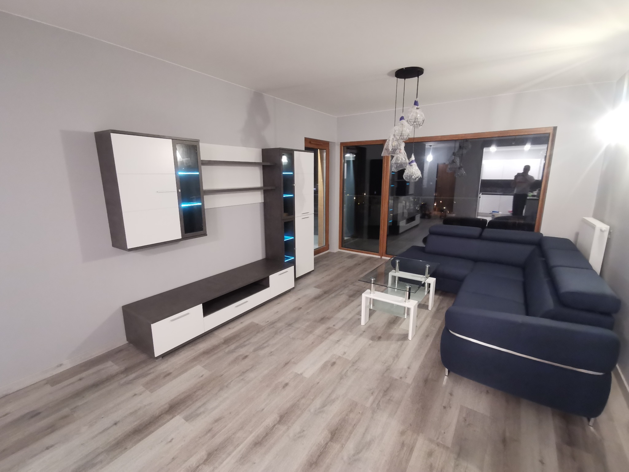 Nowoczesny salon z szarymi ścianami, meblami w kolorach bieli i grafitu, narożną sofą w odcieniach granatu oraz przeszklonymi drzwiami balkonowymi.