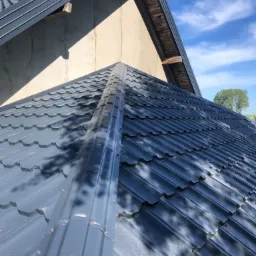 Szary dach z blachy trapezowej z widoczną kalenicą, fragmentem ściany szczytowej i niebieskim niebem w tle.