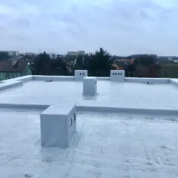 Widok z góry na płaski dach pokryty jasną membraną, z widocznymi obróbkami blacharskimi kominów i krawędzi dachu na tle zabudowy miejskiej w pochmurny dzień.